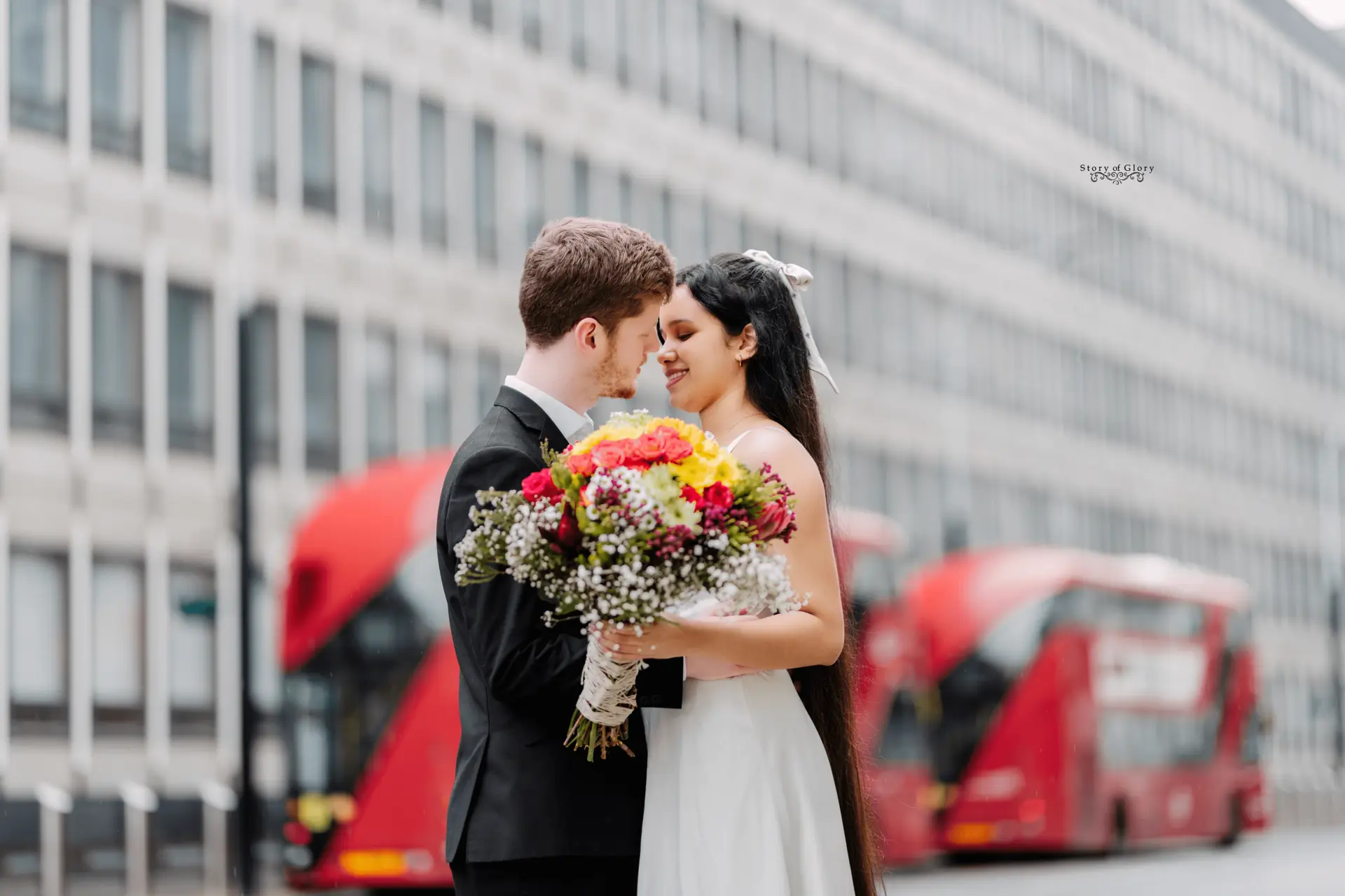 storyofglory Newlywed couple embracing with a vibrant flower bouquet in front of red double-decker buses in central London